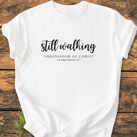 “Still Walking. 2 Corinthians 5:7 - Vintage Wash Tee” featuring black script on a white T-shirt, laid flat on a wooden surface, highlighting its relaxed fit and durable construction.