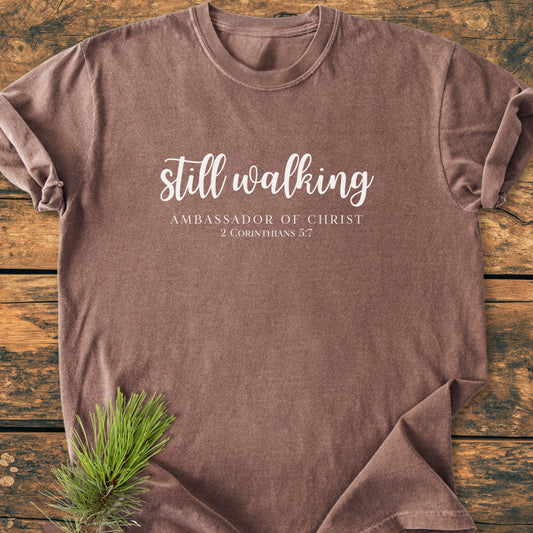 Still Walking. 2 Corinthians 5:7 - Vintage Wash Tee featuring “still walking” and “AMBASSADOR OF CHRIST” print, laid flat on a wooden surface with a pine sprig accent.