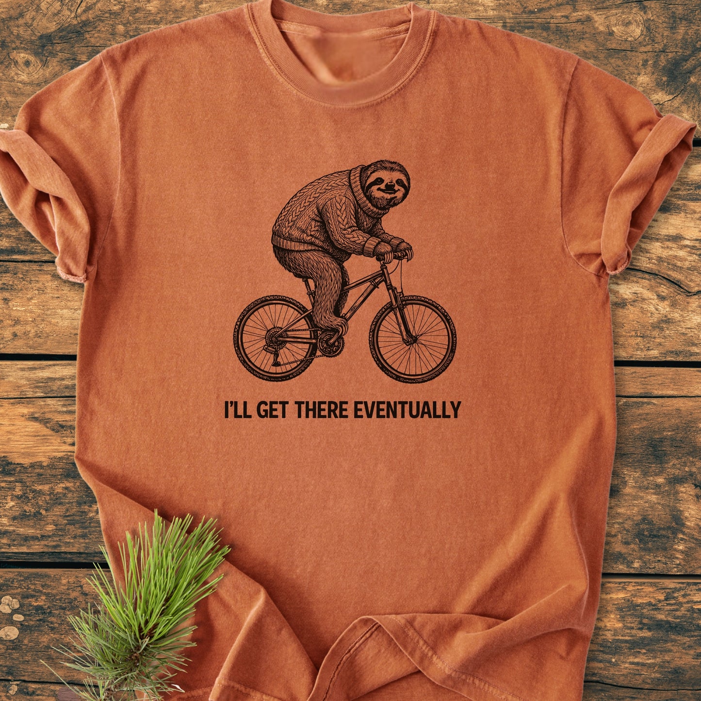 I'll Get There Eventually - Vintage Wash Tee features a sloth on a bicycle print on a washed-out orange T-shirt, displayed folded on wood, with a pine sprig beside it.