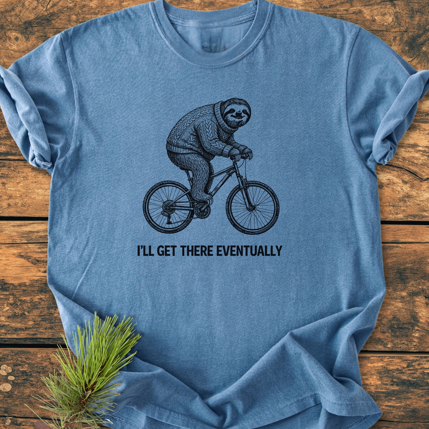 Heather-blue I'll Get There Eventually - Vintage Wash Tee featuring a sloth on a bicycle in a sweater, laid on a wooden surface with a pine sprig, highlighting comfort and durability.