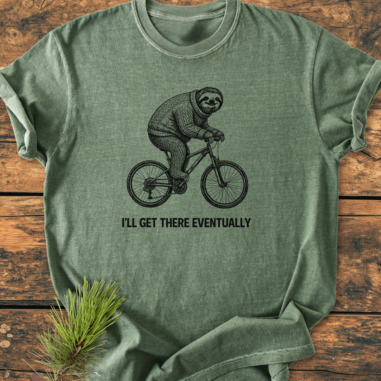 I'll Get There Eventually - Vintage Wash Tee: Heather-green T-shirt featuring a sloth on a bike, wearing a sweater, with the phrase beneath. Laid flat on wood with a pine sprig accent.