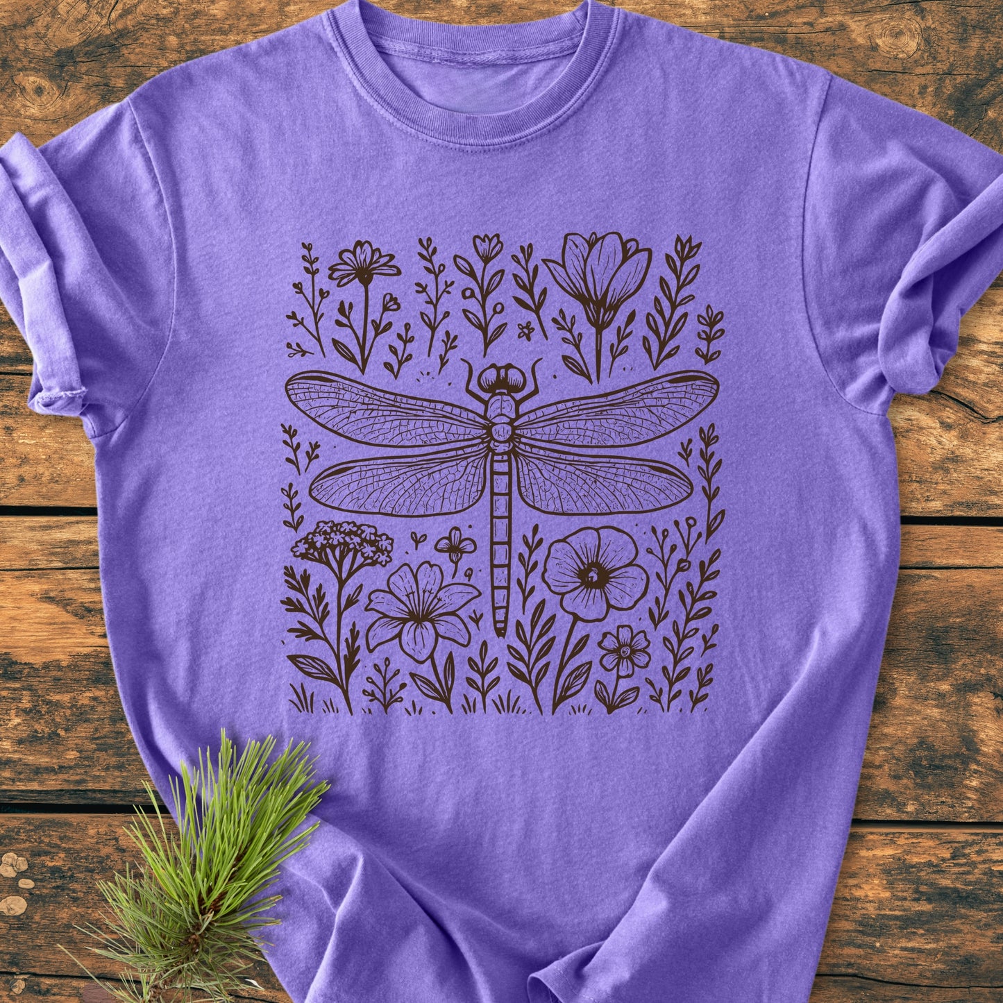 The Dragonfly - Vintage Heavyweight Tee features a hand-drawn dragonfly and wildflowers motif on a lavender-purple T-shirt, laid on a wooden surface with a pine sprig accent.
