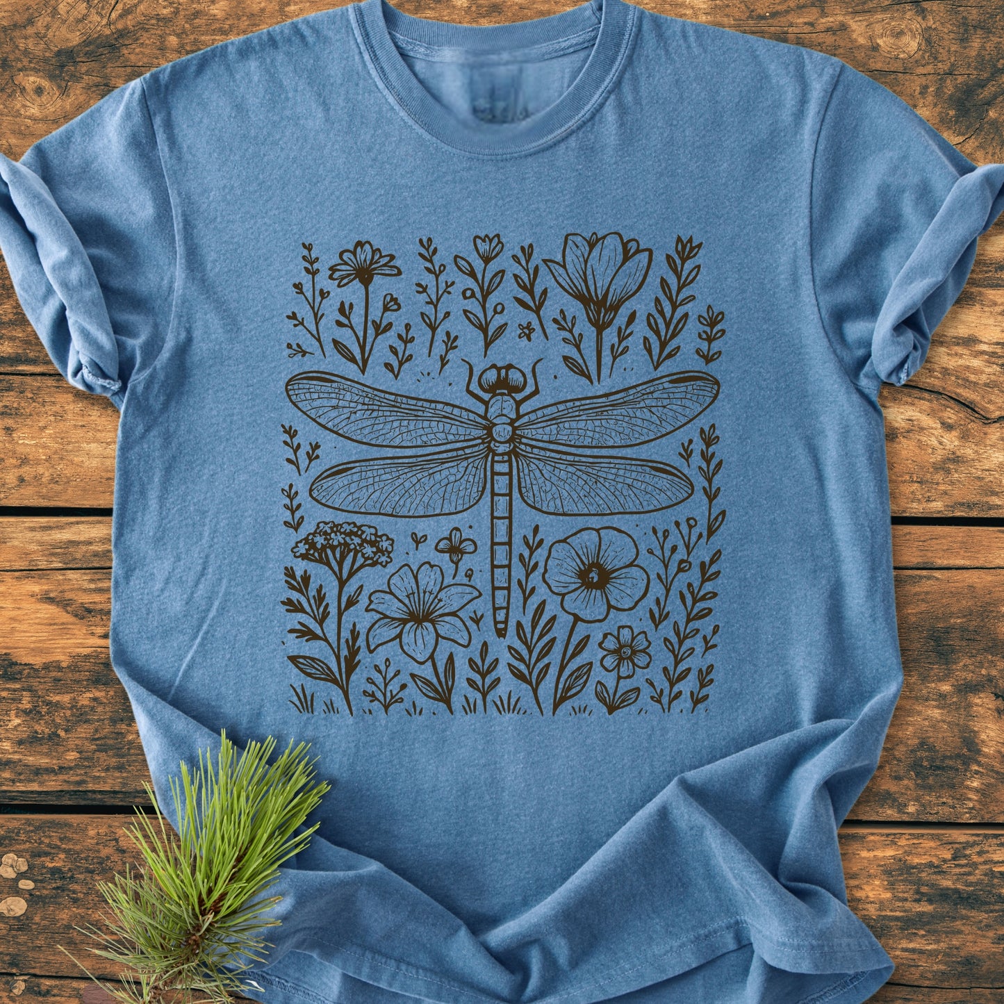 Dragonfly - Vintage Heavyweight Tee features a detailed dragonfly and wildflowers in black line-art on a heathered blue T-shirt, presented with a rustic touch on a wooden background.