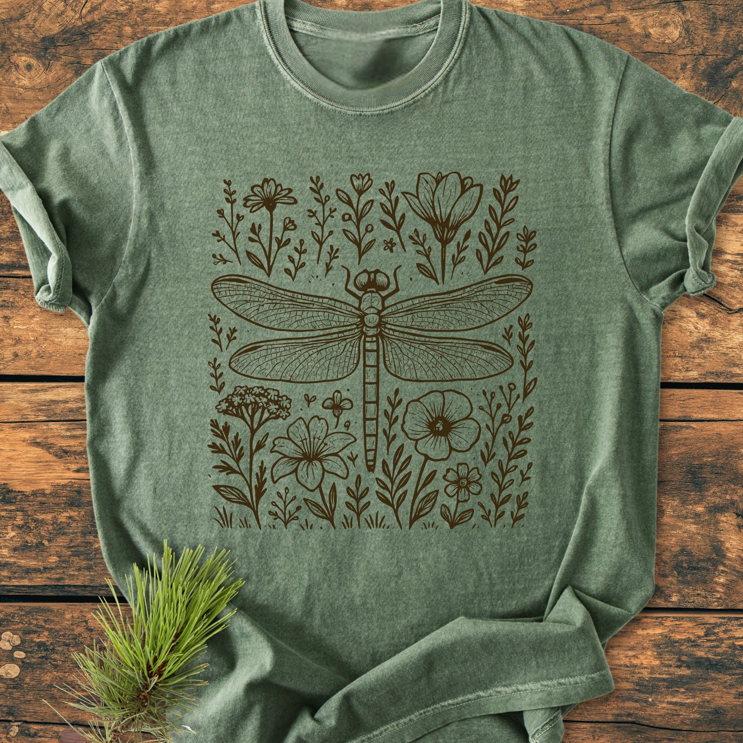 Dragonfly - Vintage Heavyweight Tee displayed on wooden surface, features a dragonfly illustration with wildflowers. Styled with a pine branch for an outdoorsy touch, emphasizing its relaxed, durable design.
