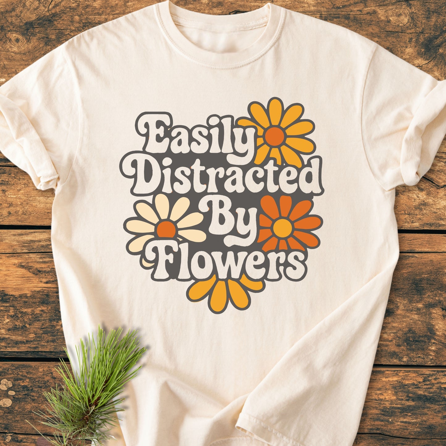 Easily Distracted By Flowers - Vintage Heavyweight Tee features retro-style lettering and daisy-like blooms on a cream T-shirt, laid flat with a pine sprig accent on a wooden surface.