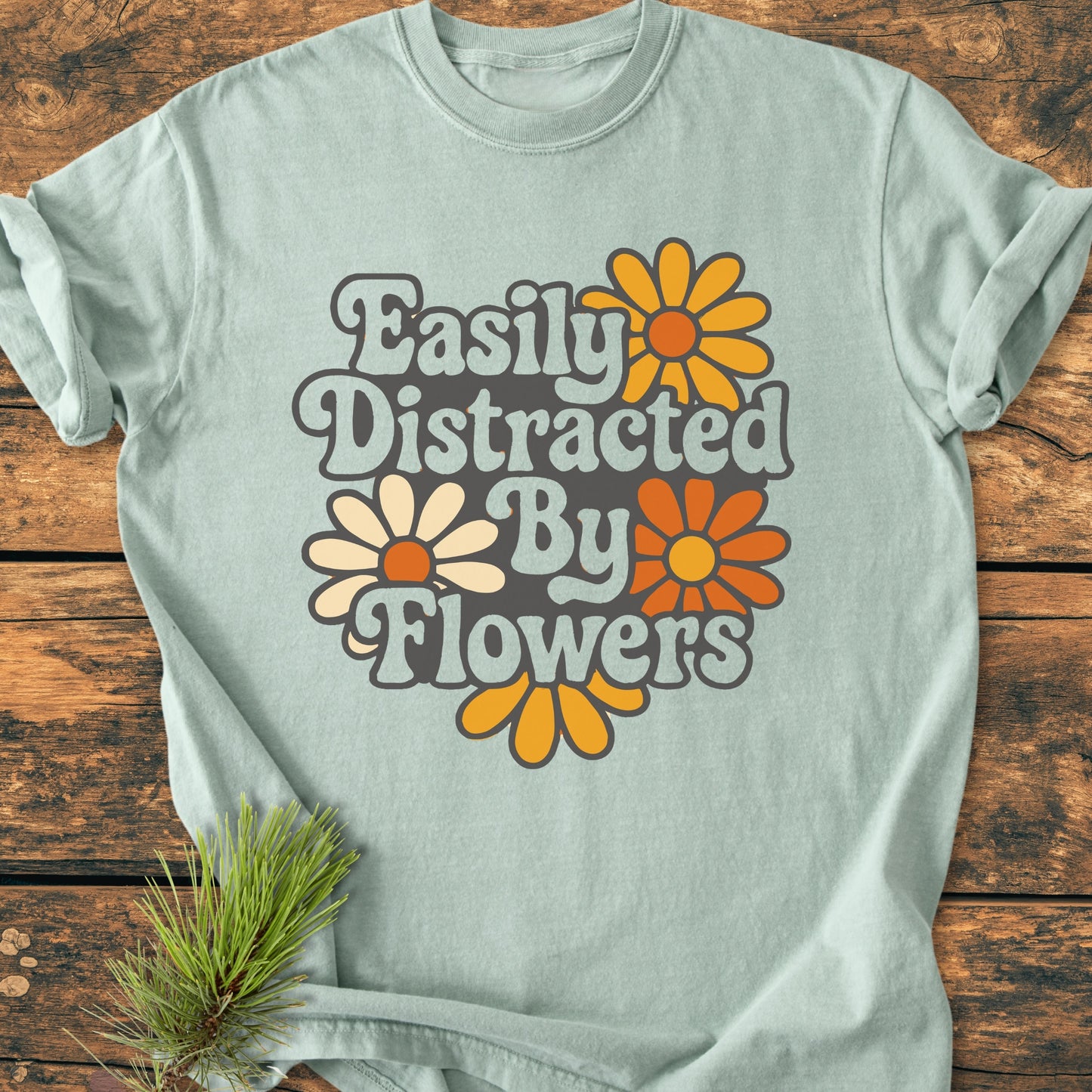 Easily Distracted By Flowers - Vintage Heavyweight Tee features 1970s-inspired lettering and daisy graphics on a mint T-shirt, laid on wood, styled with a pine sprig for a cozy, retro look.