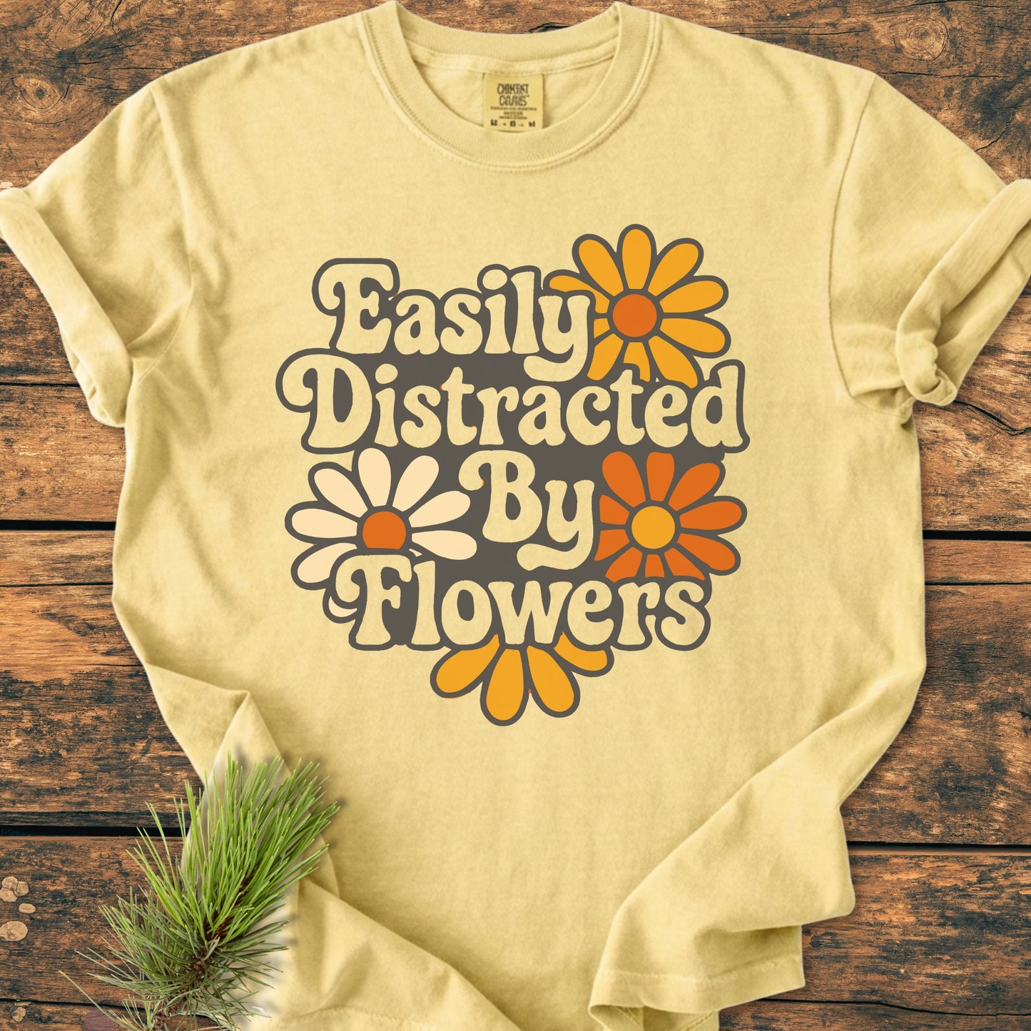 Easily Distracted By Flowers - Vintage Heavyweight Tee featuring a groovy '70s font and daisy blooms, laid flat on wood, showcasing its soft, relaxed, garment-dyed fabric.