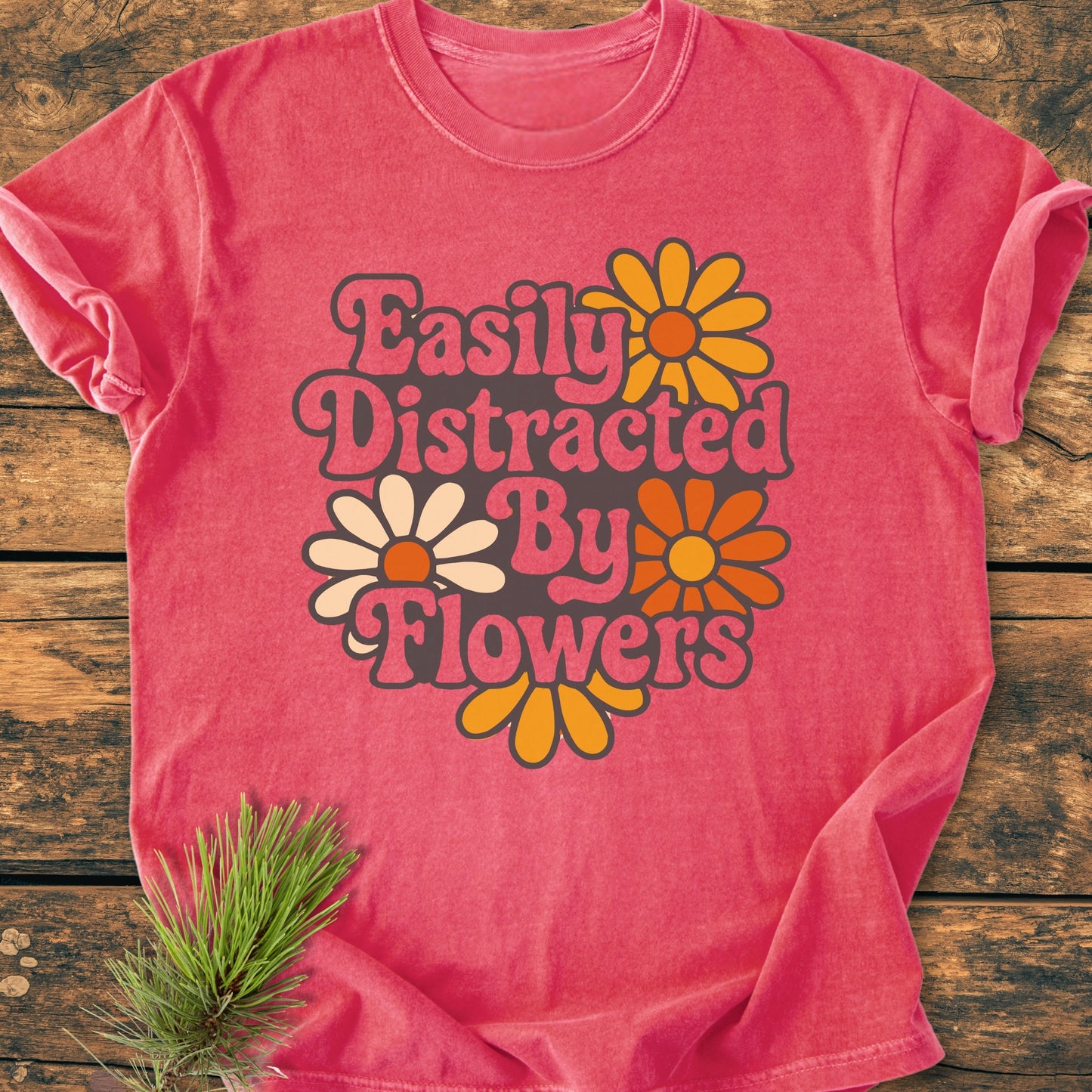 Easily Distracted By Flowers - Vintage Heavyweight Tee features a playful design of stylized daisies and retro lettering on a heathered pink T-shirt, styled with a pine sprig on rustic wood.