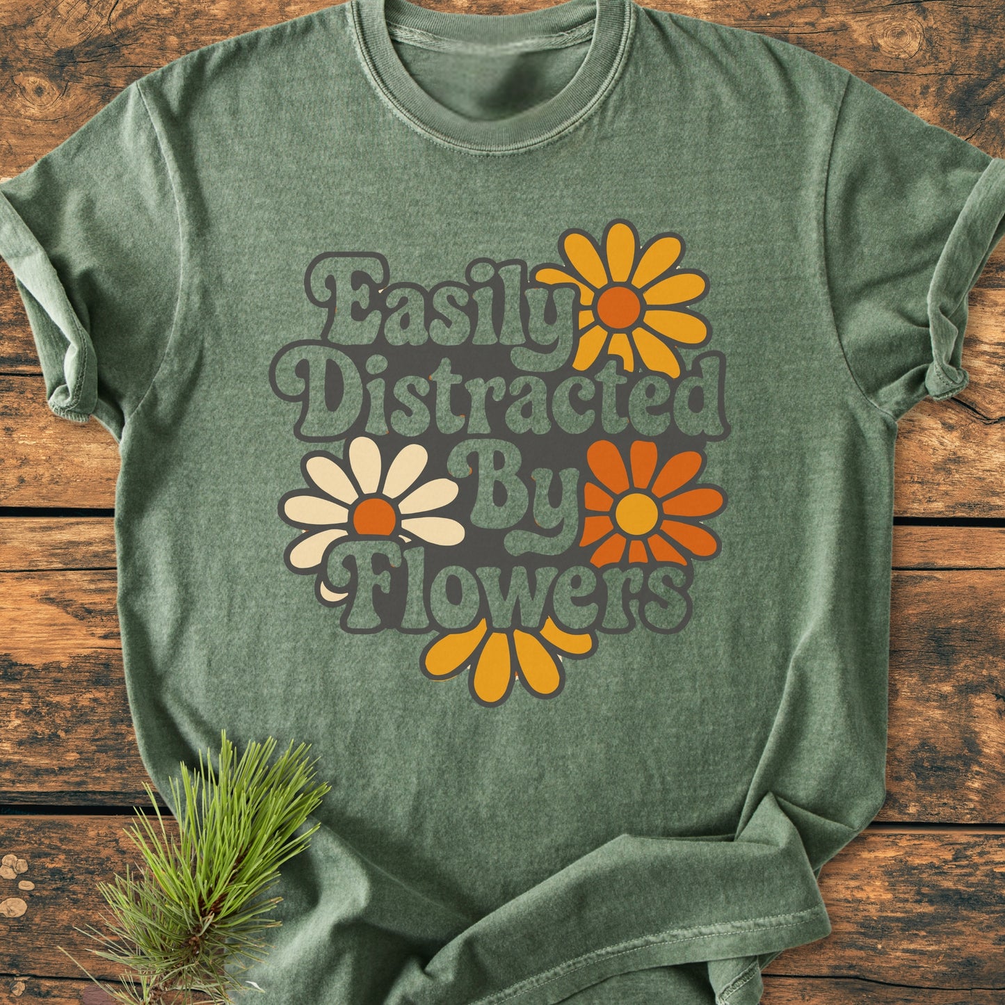 Easily Distracted By Flowers - Vintage Heavyweight Tee features a retro graphic with daisies and slogan, laid on wooden background, showcasing its heather-green hue and relaxed, cozy fit.