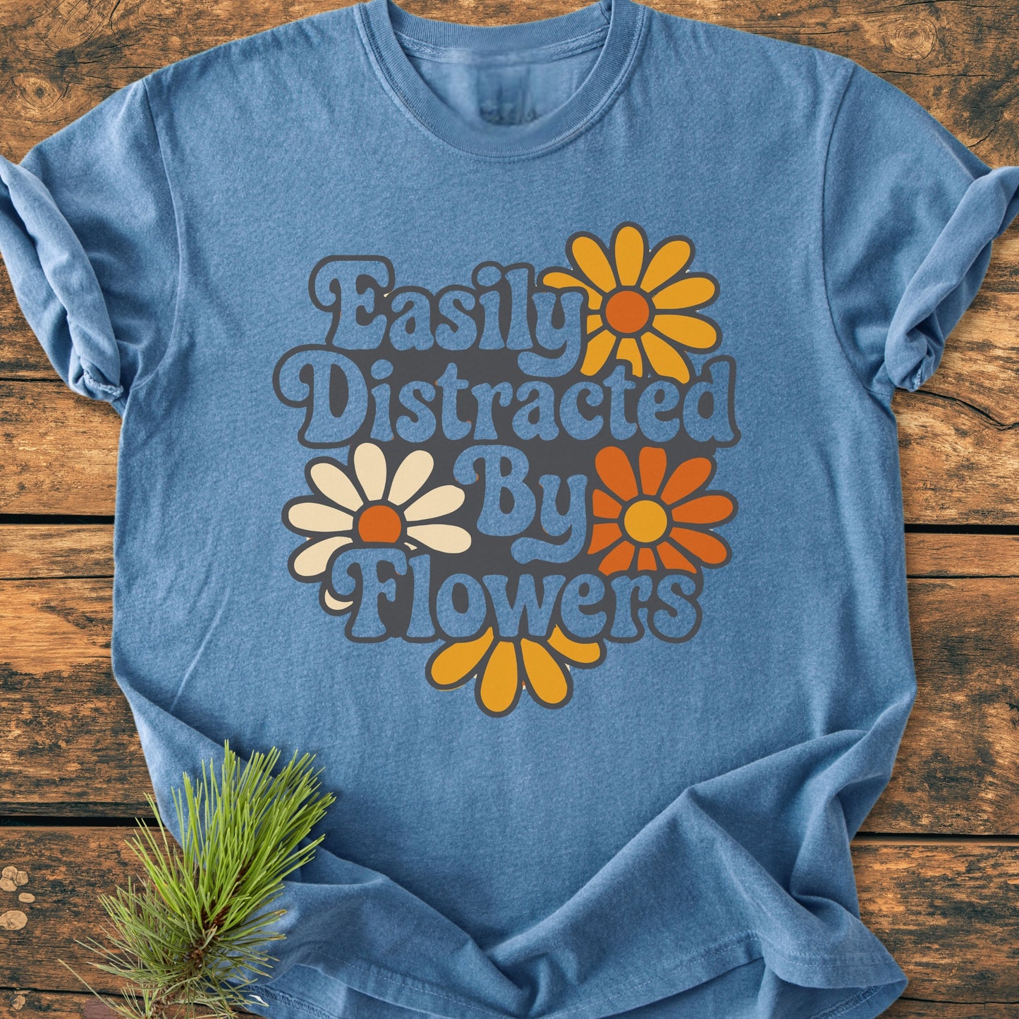 Easily Distracted By Flowers - Vintage Heavyweight Tee in heather-blue, featuring retro lettering and daisy prints. Displayed on wood with a pine sprig, highlighting its cozy, relaxed fit.