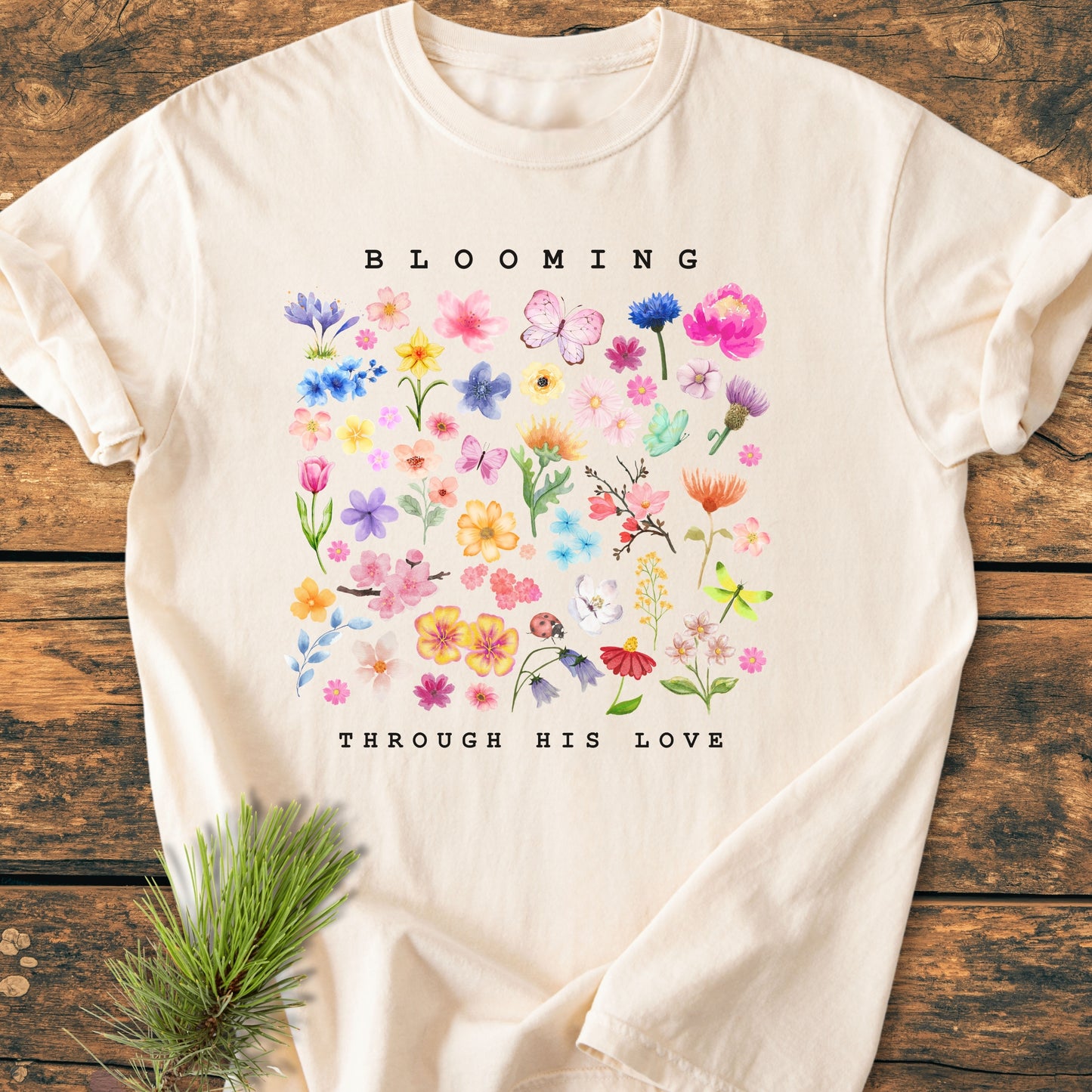 Blooming Through His Love - Vintage Wash Tee features a watercolor spring flower grid with butterflies, a ladybug, and dragonfly, and the phrase BLOOMING THROUGH HIS LOVE on a soft, relaxed-fit, garment-dyed cotton.