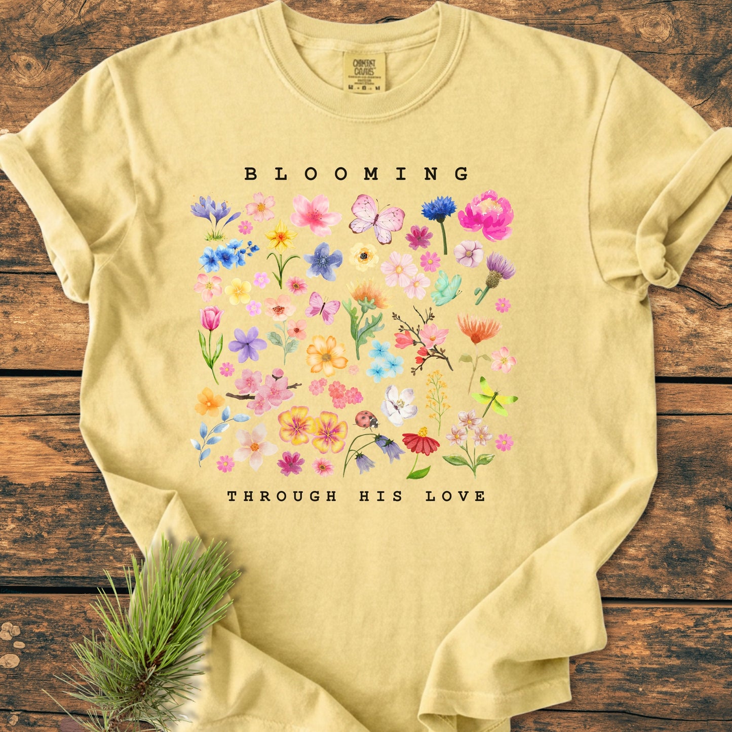 Blooming Through His Love - Vintage Wash Tee features a grid of watercolor flowers, butterflies, and a ladybug, with BLOOMING and THROUGH HIS LOVE text, displayed on a wooden surface.