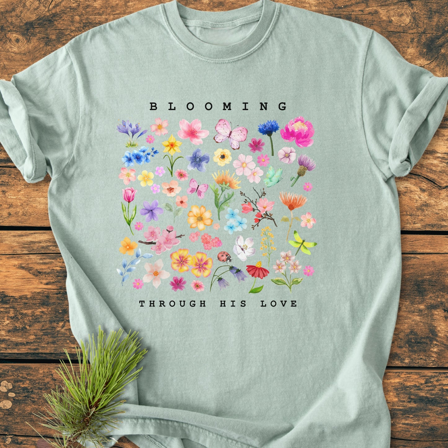 Blooming Through His Love - Vintage Wash Tee: Light sage-green T-shirt featuring a pastel floral grid with butterflies, ladybugs, and inspirational text, laid flat on a wooden surface with a pine sprig beside it.