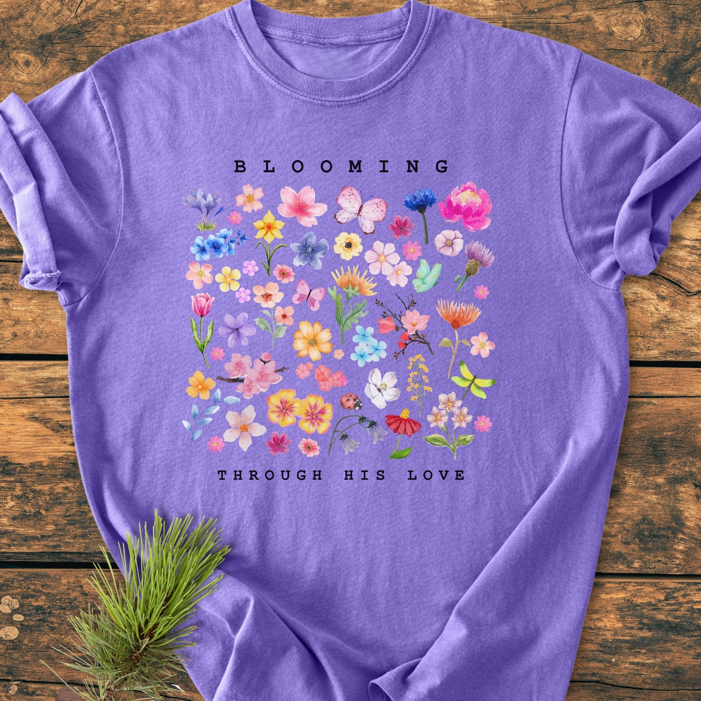 Blooming Through His Love - Vintage Wash Tee featuring a watercolor floral and butterfly design, laid on wood with a pine sprig. Soft lavender crew-neck, showcasing Comfort Colors' durable, relaxed-fit garment.