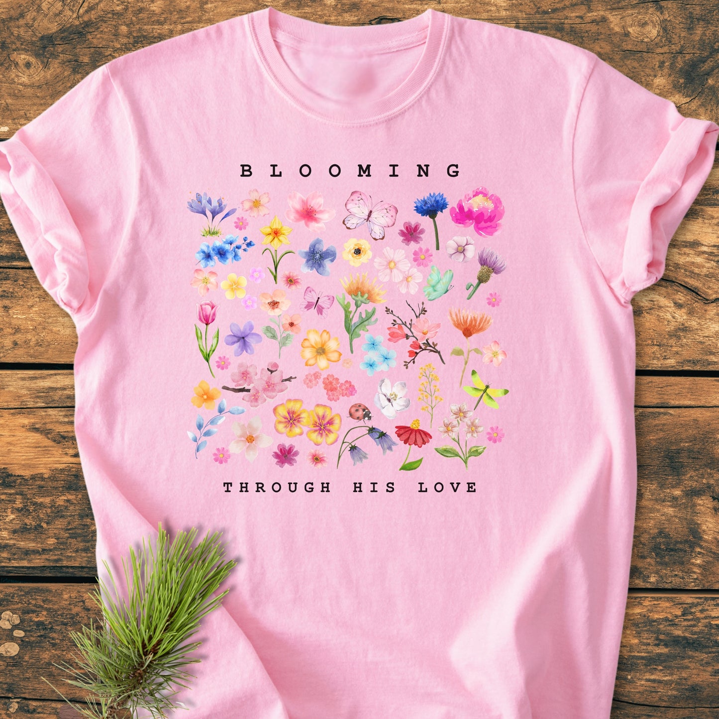 Blooming Through His Love - Vintage Wash Tee featuring a light-pink hue with colorful watercolor-style flowers, insects, and bold text. Laid on wood with a pine sprig accent.