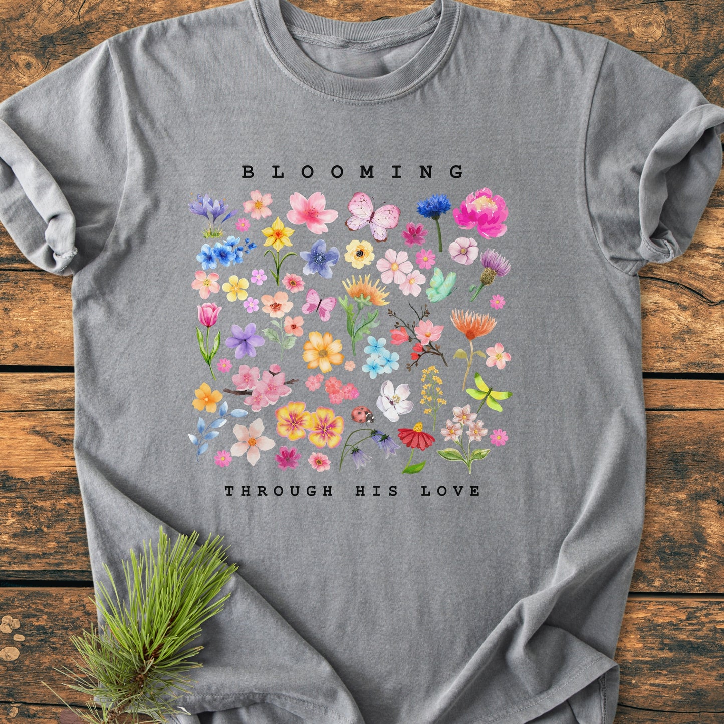 Blooming Through His Love - Vintage Wash Tee featuring watercolor-style flowers and butterflies, BLOOMING text above, on a heather-gray background with a cozy, relaxed fit, styled with a fir branch.
