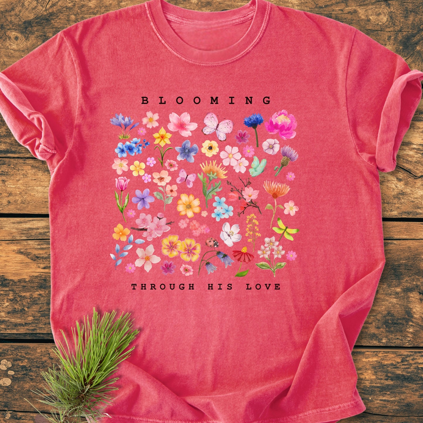 Blooming Through His Love - Vintage Wash Tee features a colorful floral and insect design with BLOOMING THROUGH HIS LOVE text, laid on a wooden surface, showcasing its relaxed fit and durability.