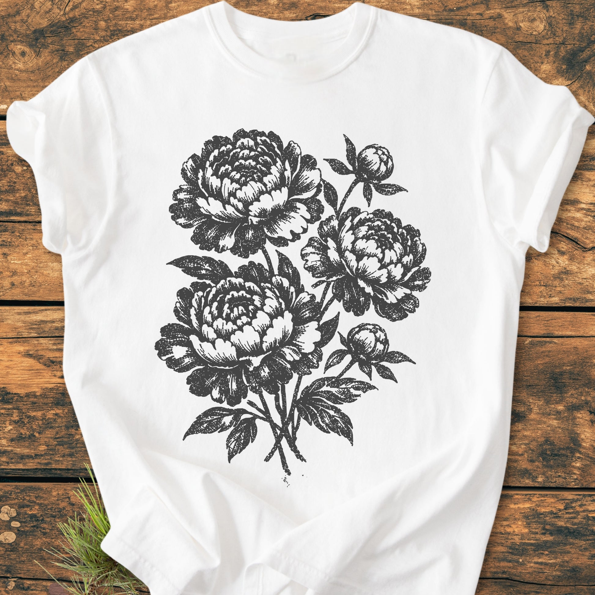 Peony Bouquet - Vintage Heavyweight Tee features a black woodcut-style floral graphic of peonies printed on a white crew-neck T-shirt, laid flat on a rustic wooden surface, highlighting its relaxed, durable design.