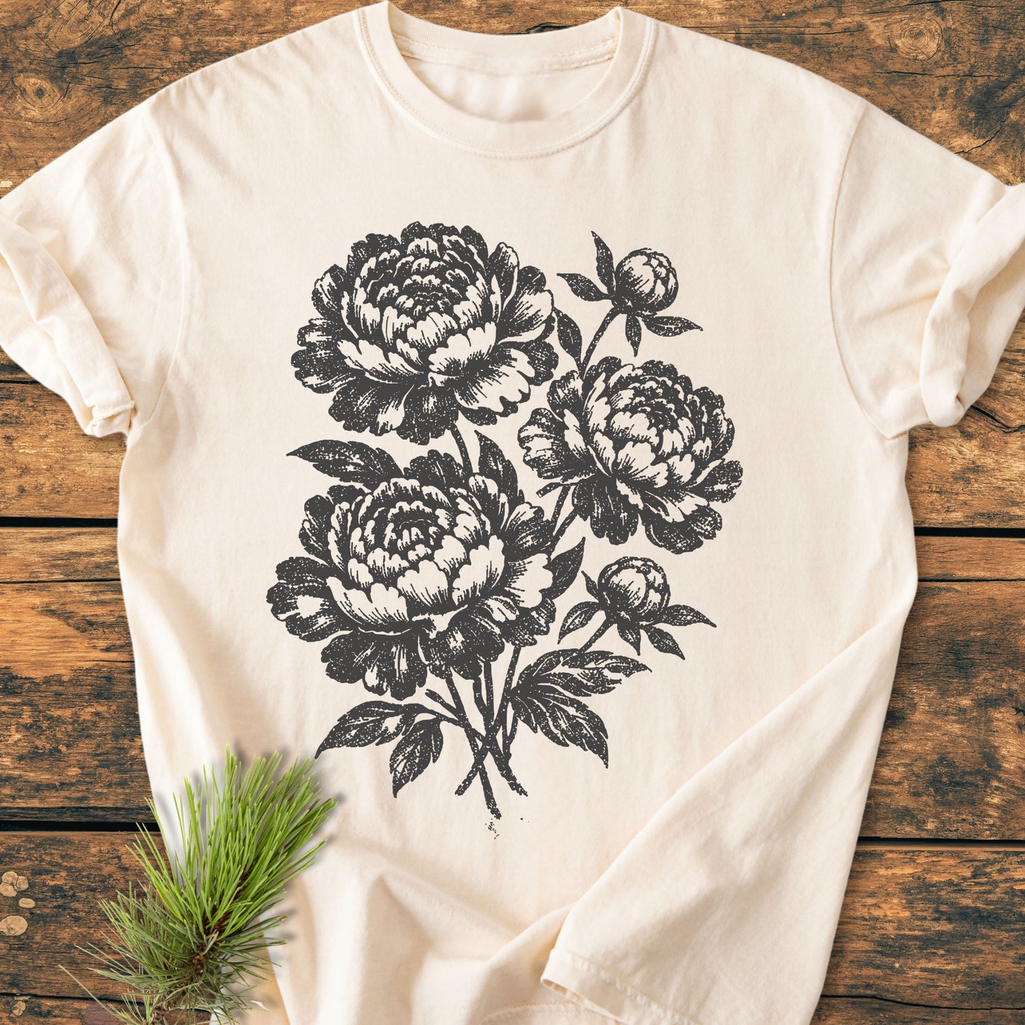 Peony Bouquet - Vintage Heavyweight Tee features a black line-art of peony blossoms on a cream T-shirt, laid flat on wood. Accentuated with an evergreen sprig for a natural touch.