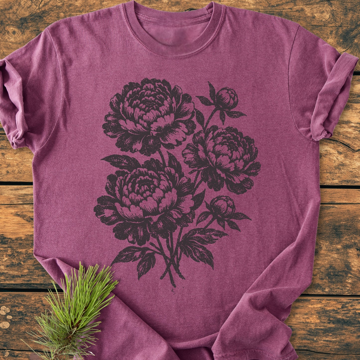 Peony Bouquet - Vintage Heavyweight Tee, a heathered plum-purple T-shirt with black botanical peony illustration, laid flat with casually rolled sleeves on a wooden surface, featuring a small pine sprig.