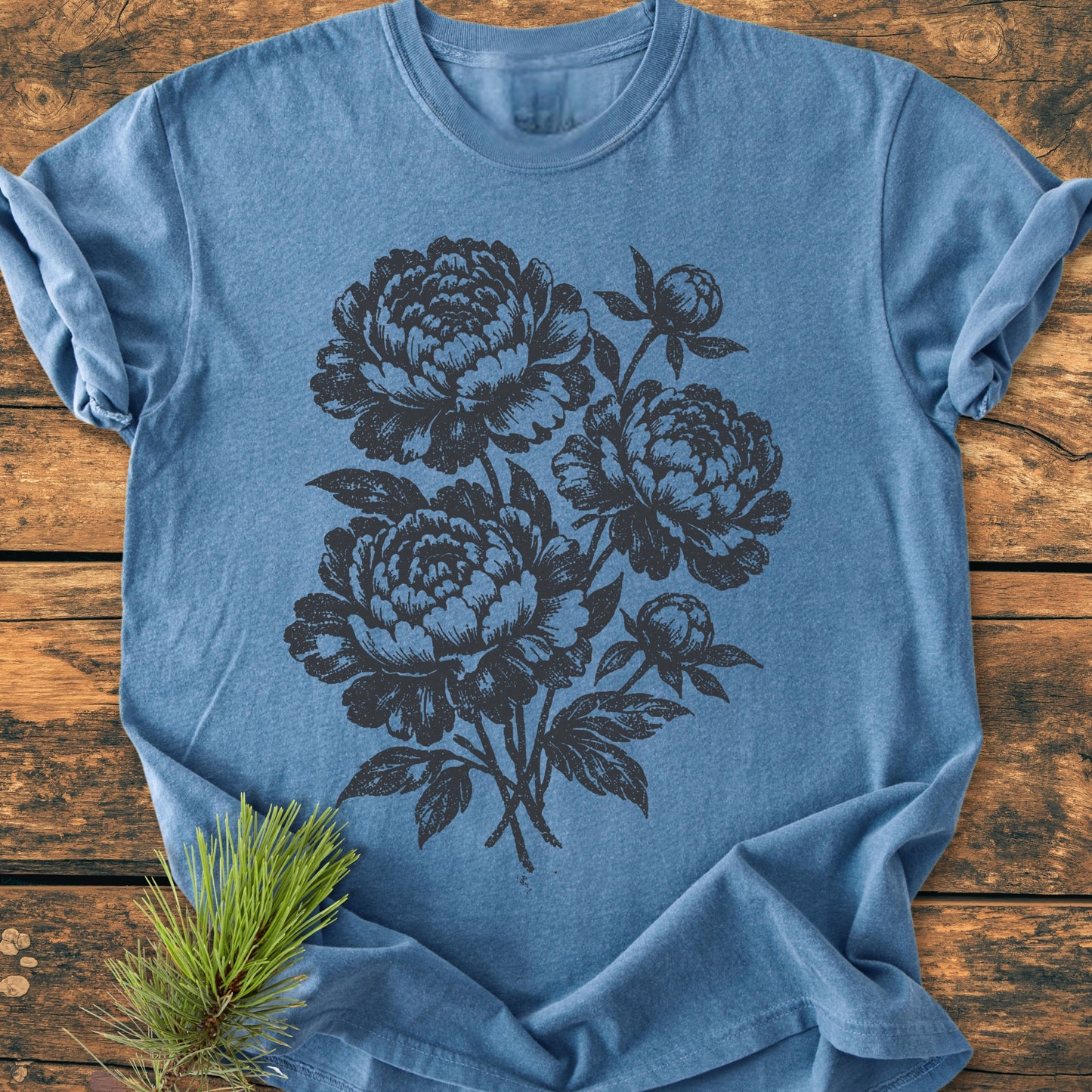 Peony Bouquet - Vintage Heavyweight Tee, featuring black-screen-printed peony-style flowers, displayed on a rustic wooden surface. Includes three large blossoms, two buds, and leaves; a small pine sprig accents the design.