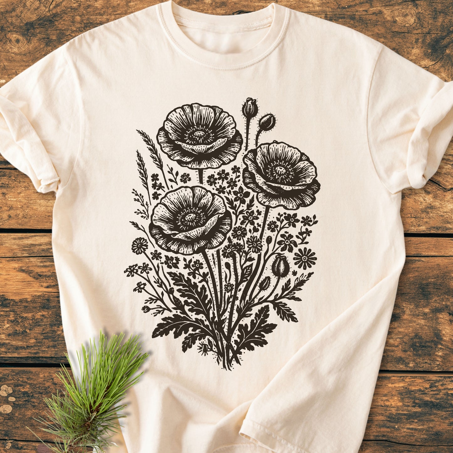 Wildflower Bouquet - Vintage Heavyweight Tee features a detailed black line-art botanical design with large poppy blooms and wildflowers on a light T-shirt, displayed on a wooden surface with a pine sprig.