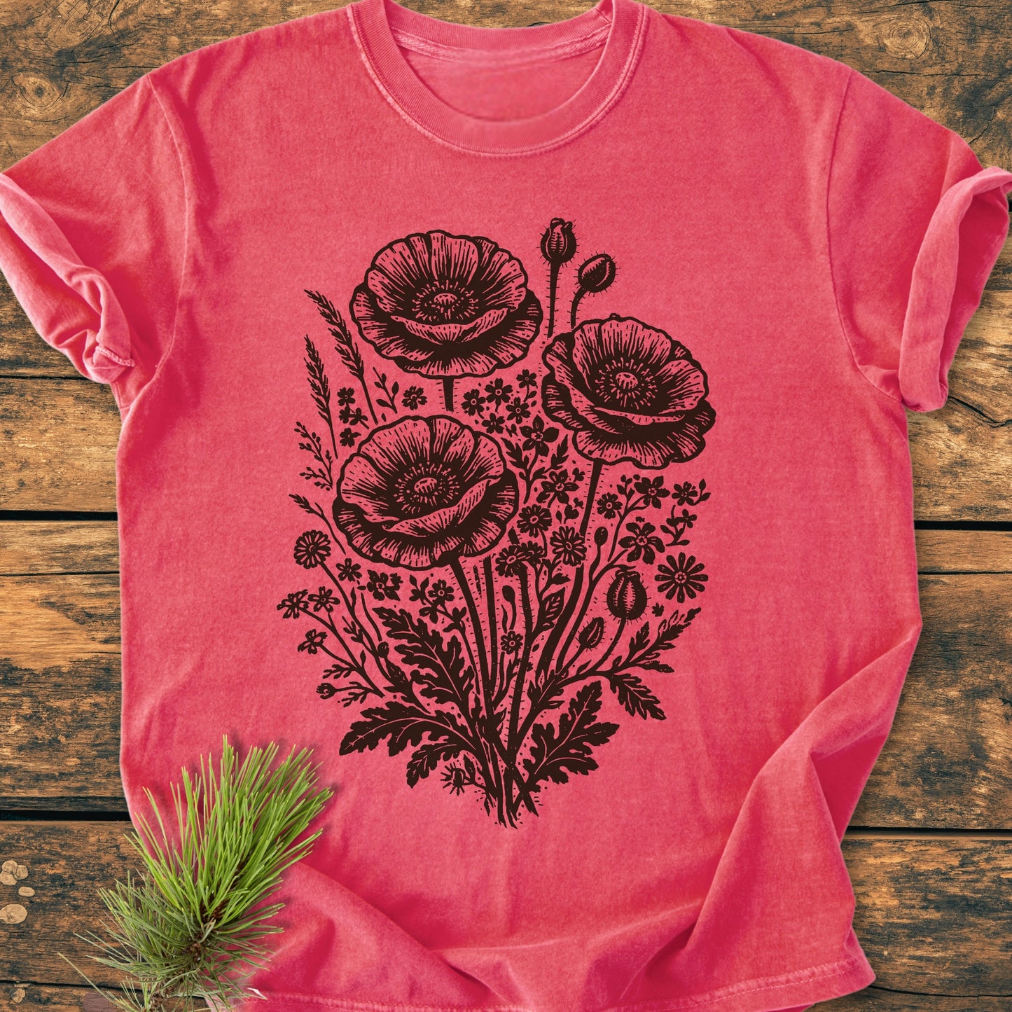 Wildflower Bouquet - Vintage Heavyweight Tee features a black botanical poppy illustration on a coral-pink T-shirt, displayed on wood. A pine twig accentuates the design's natural, cozy aesthetic.