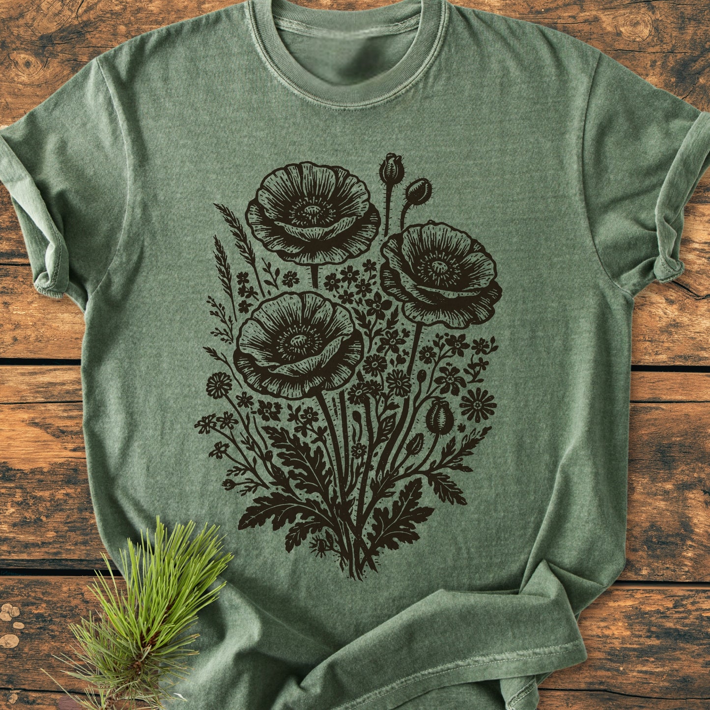 Wildflower Bouquet - Vintage Heavyweight Tee in heather-green, features a dark, detailed wildflower design. Laid on a wooden surface, styled with a pine twig, showcasing its relaxed fit and intricate print.