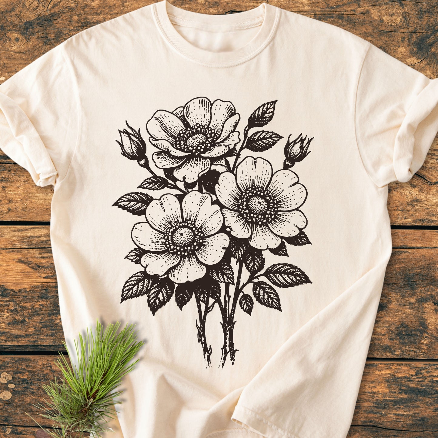 Wild Rose Bouquet - Vintage Heavyweight Tee: A light-cream, short-sleeve T-shirt with a black botanical illustration of wild-rose flowers and buds, displayed on a rustic wooden surface with a small pine branch.