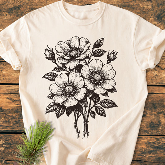 Wild Rose Bouquet - Vintage Heavyweight Tee: A light-cream, short-sleeve T-shirt with a black botanical illustration of wild-rose flowers and buds, displayed on a rustic wooden surface with a small pine branch.
