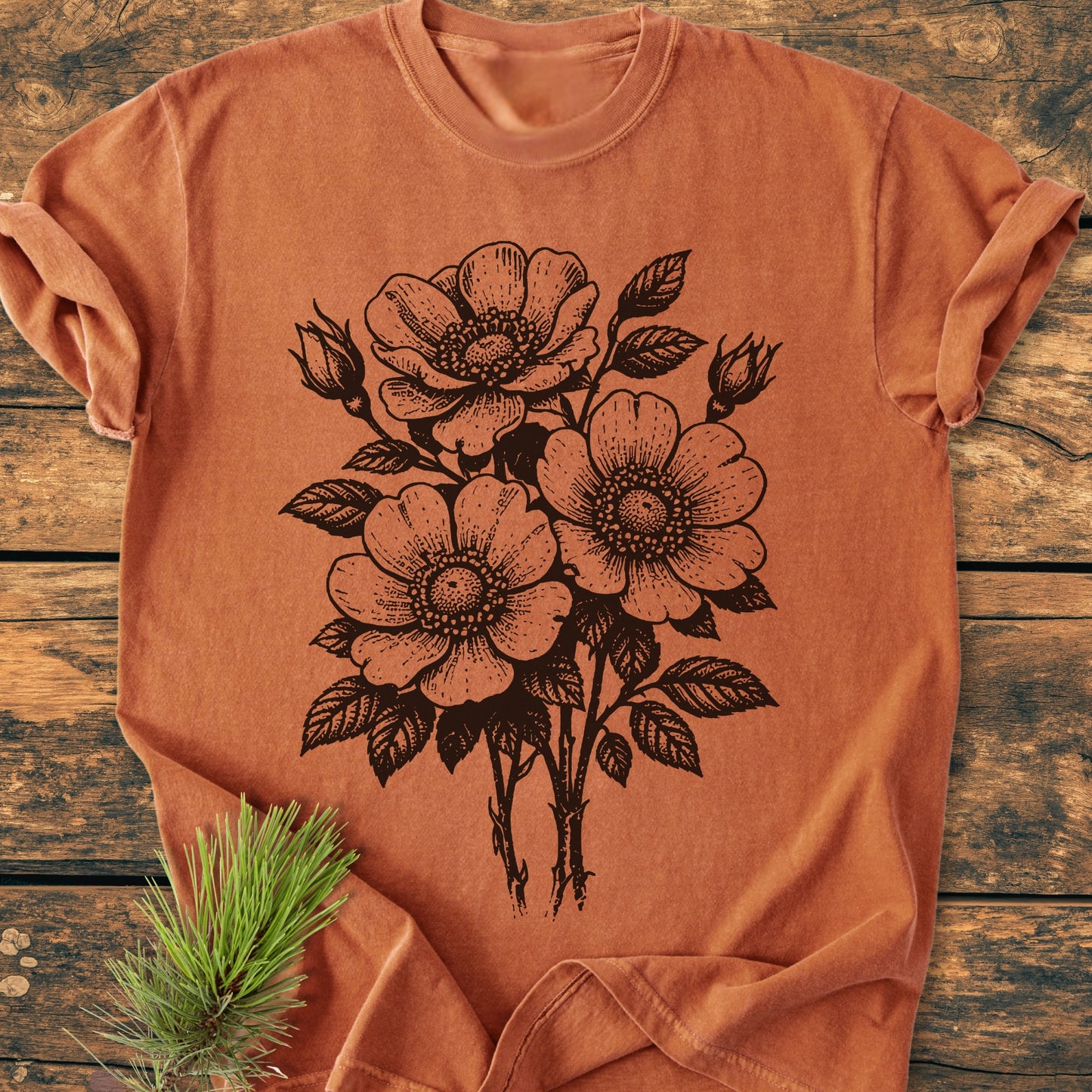 Wild Rose Bouquet - Vintage Heavyweight Tee in burnt-orange, featuring black botanical illustration of wild roses. Laid on wooden surface with pine sprig. Made from soft, garment-dyed, 100% ring-spun cotton.