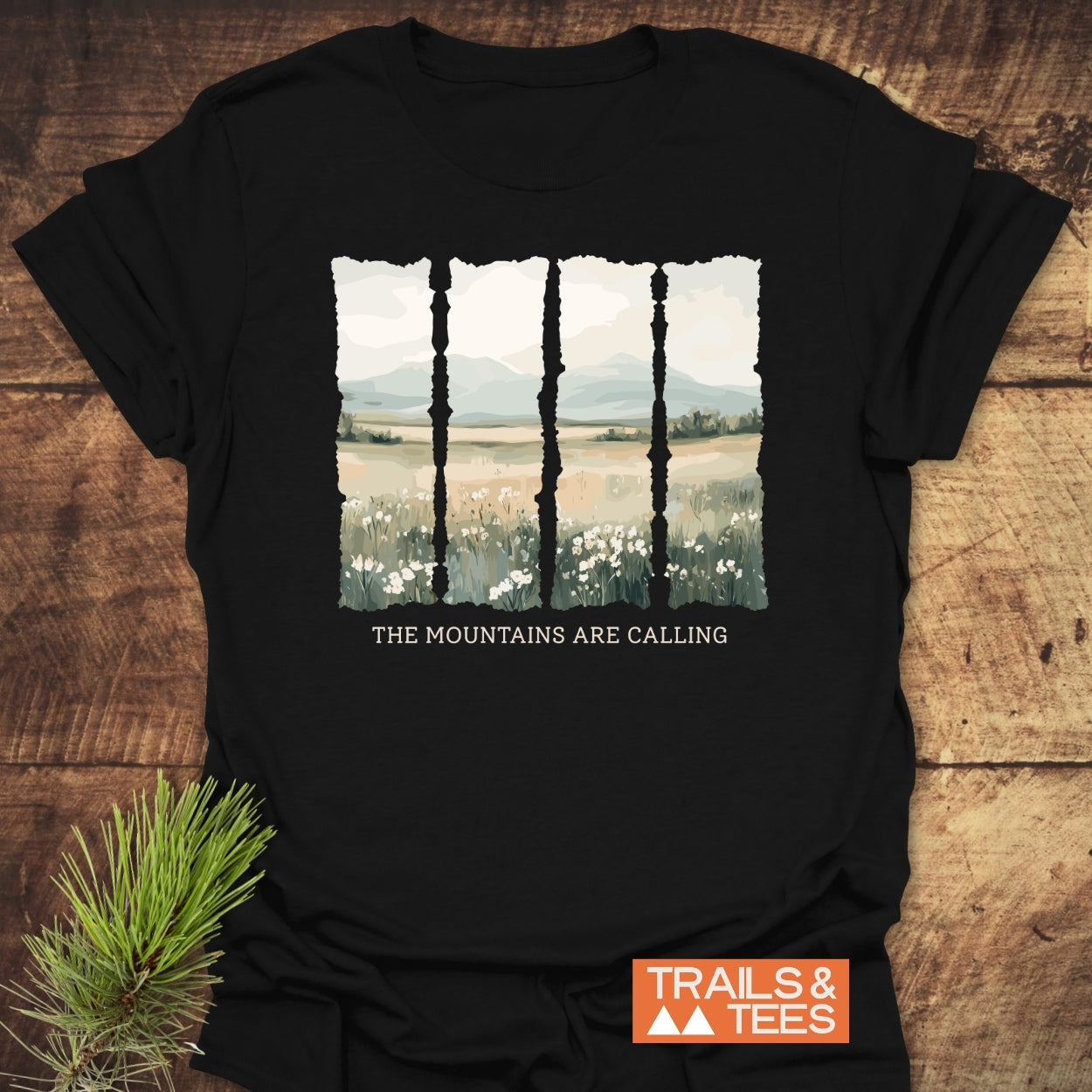 Mountains Are Calling T-Shirt featuring a stylized landscape with wildflowers, hills, and mountains. Text beneath reads “THE MOUNTAINS ARE CALLING.” Includes a small “TRAILS & TEES” logo.