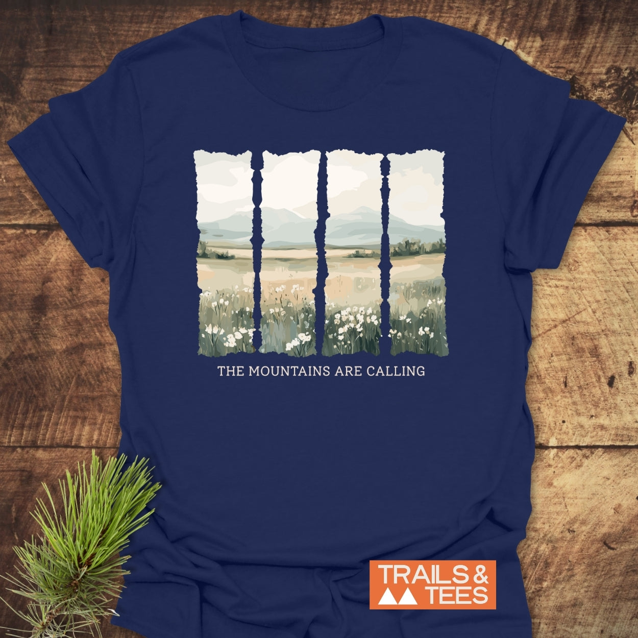Mountains Are Calling T-Shirt featuring a stylized landscape in torn-edge panels, depicting wildflowers, fields, and mountains. Includes slogan and “TRAILS & TEES” logo, laid on a wooden tabletop with pine sprig.