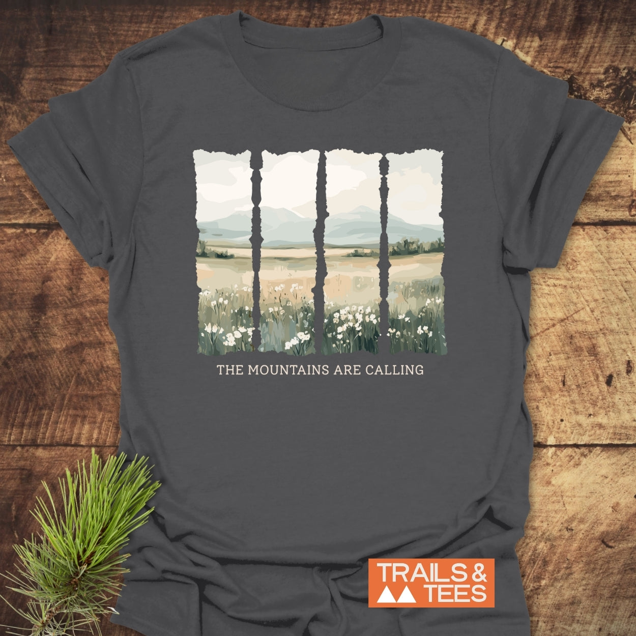 Mountains Are Calling T-Shirt displaying a painterly landscape in five panels, featuring white wildflowers, meadow, and soft-blue mountains. Beneath the artwork, the slogan reads “THE MOUNTAINS ARE CALLING.”