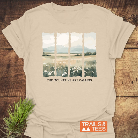 Mountains Are Calling T-Shirt on wooden surface, featuring a landscape with wildflowers and mountains. Includes slogan, THE MOUNTAINS ARE CALLING, TRAILS & TEES branding, and pine sprig accessory.
