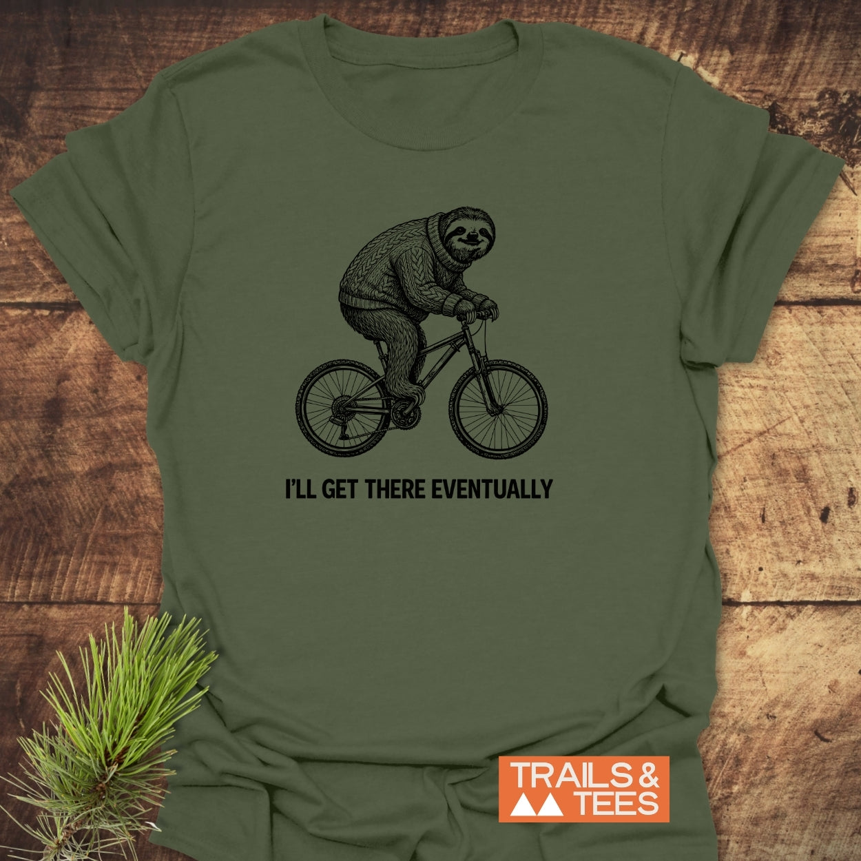 I'll Get There Eventually T-Shirt featuring a hand-drawn sloth cycling in a sweater. Crafted from soft, ring-spun cotton, it embodies outdoor adventure. Includes 'TRAILS & TEES' logo, laid on a wooden surface.