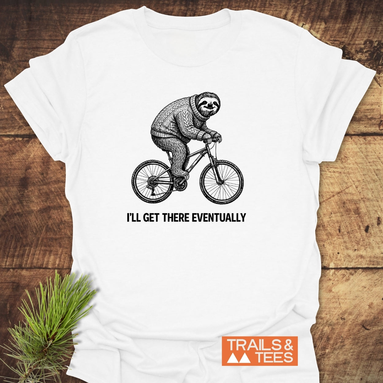 I'll Get There Eventually T-Shirt: Features a sloth in a sweater on a bike with a motivational caption, highlighting outdoor adventure spirit. Includes 'TRAILS & TEES' logo, emphasizing comfort and eco-conscious design.