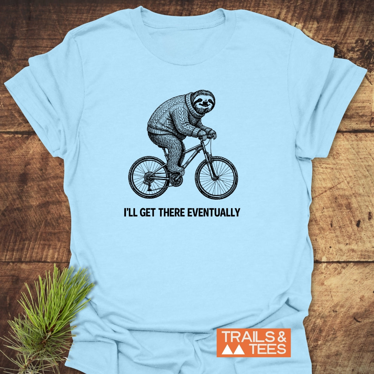 Light-blue I'll Get There Eventually T-Shirt featuring a sloth in a cable-knit sweater biking, with a motivational slogan, laid on a wooden surface, accompanied by a TRAILS & TEES logo.
