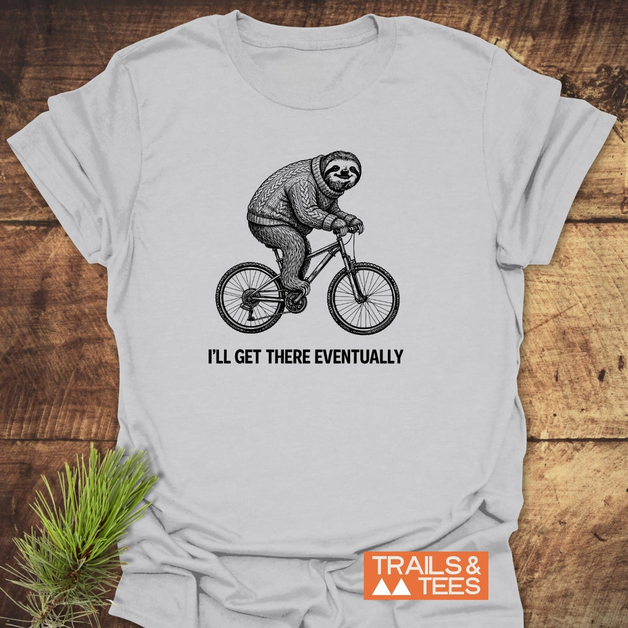I'll Get There Eventually T-Shirt featuring a sloth on a bicycle in a cozy sweater, laid flat. Includes 'TRAILS & TEES' logo, crafted from soft, ring-spun cotton for outdoor enthusiasts.