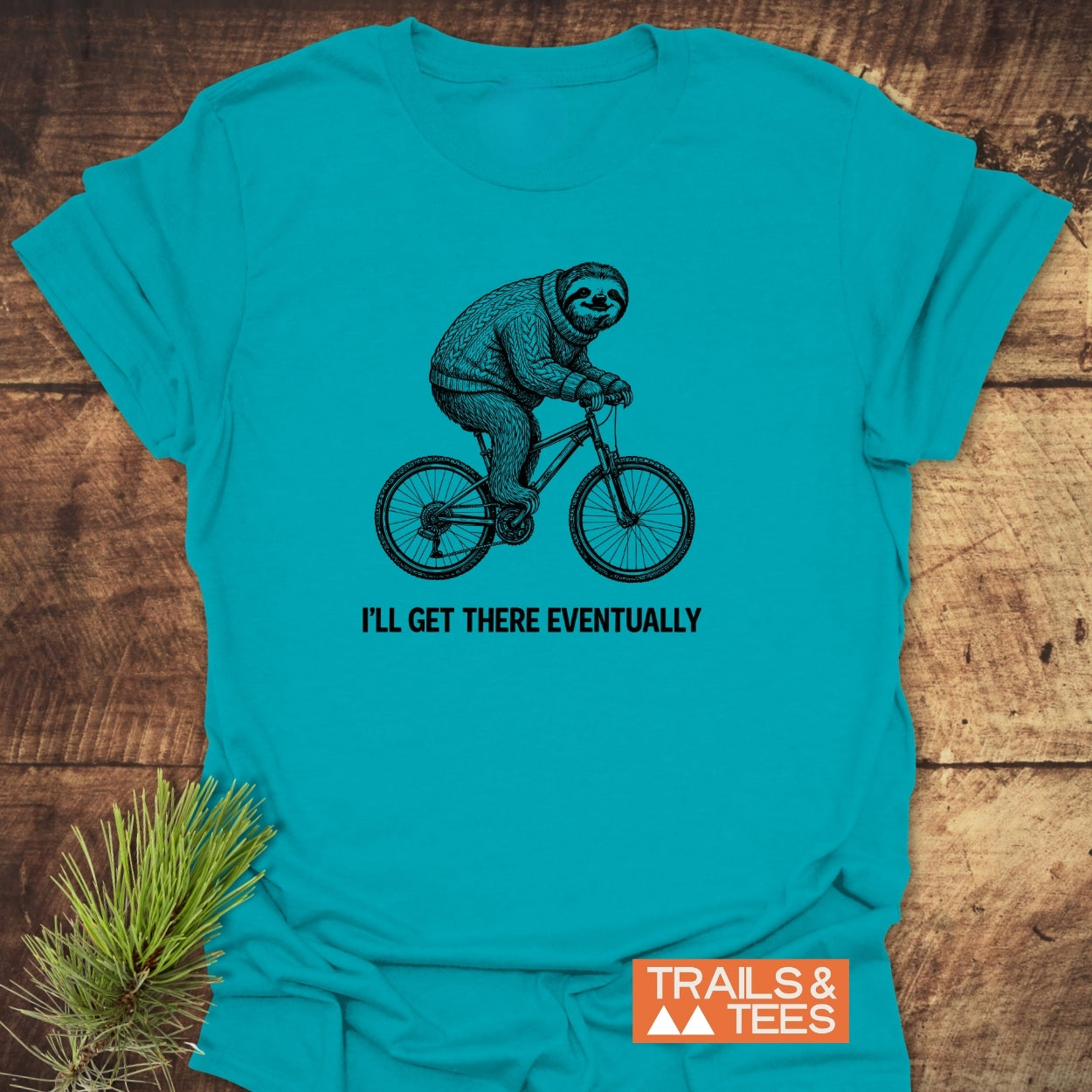 I'll Get There Eventually T-Shirt features a hand-drawn sloth on a bicycle with a slogan, crafted from soft cotton for outdoor enthusiasts, highlighted by Trails & Tees' logo and pine needles.