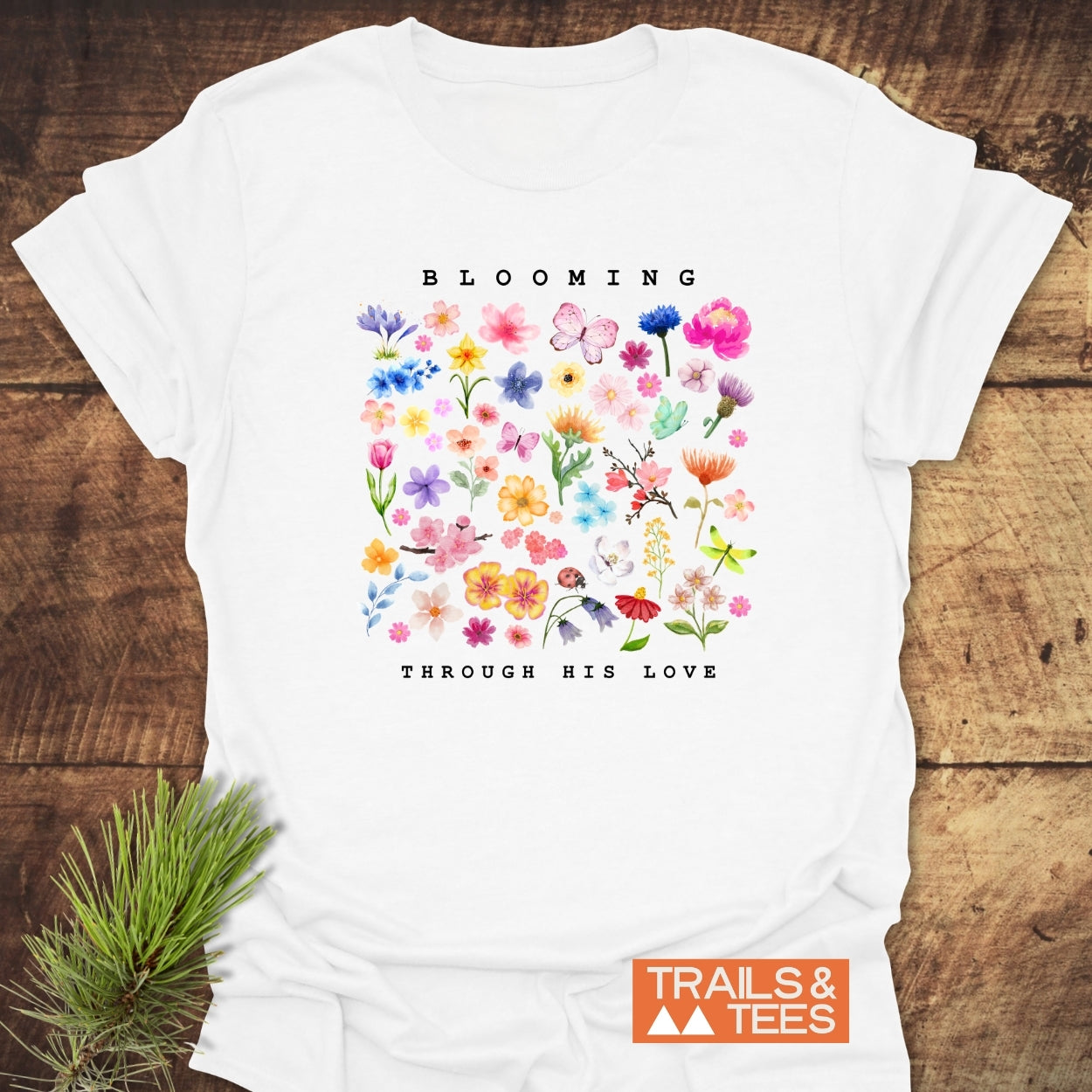 The Blooming Through His Love T-Shirt features a vivid floral and butterfly grid design with uplifting text. Crafted from soft cotton, it embodies outdoor adventure and comfort, branded by Trails & Tees.