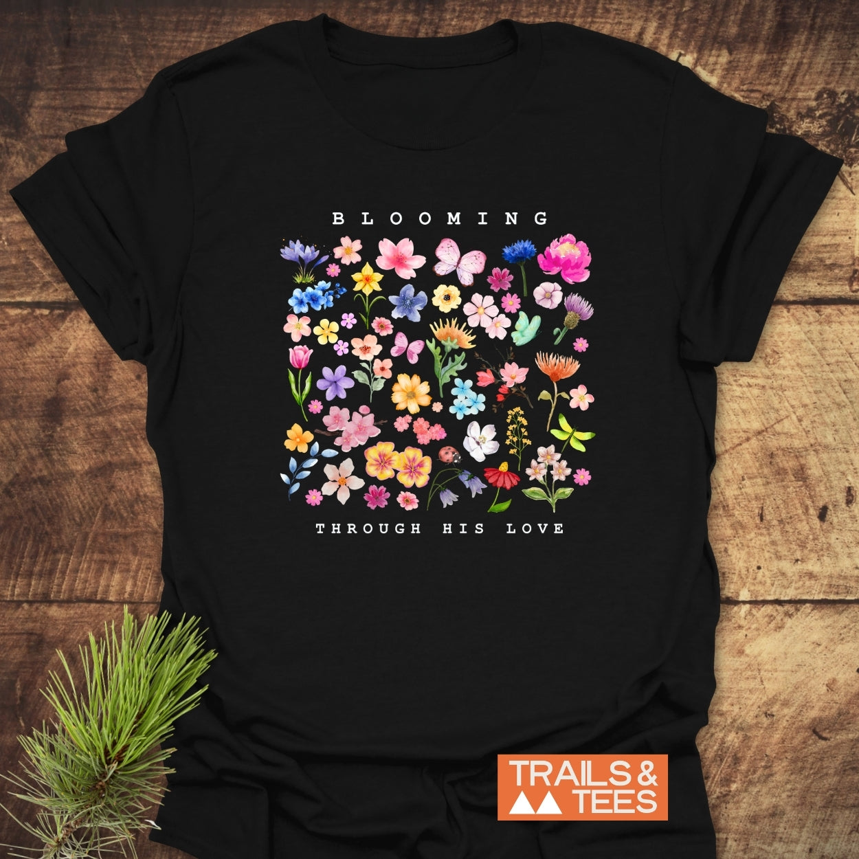 Blooming Through His Love T-Shirt features a colorful floral grid with butterflies and uplifting text on a black tee. Brand logo TRAILS & TEES is visible, laid on a wooden surface.
