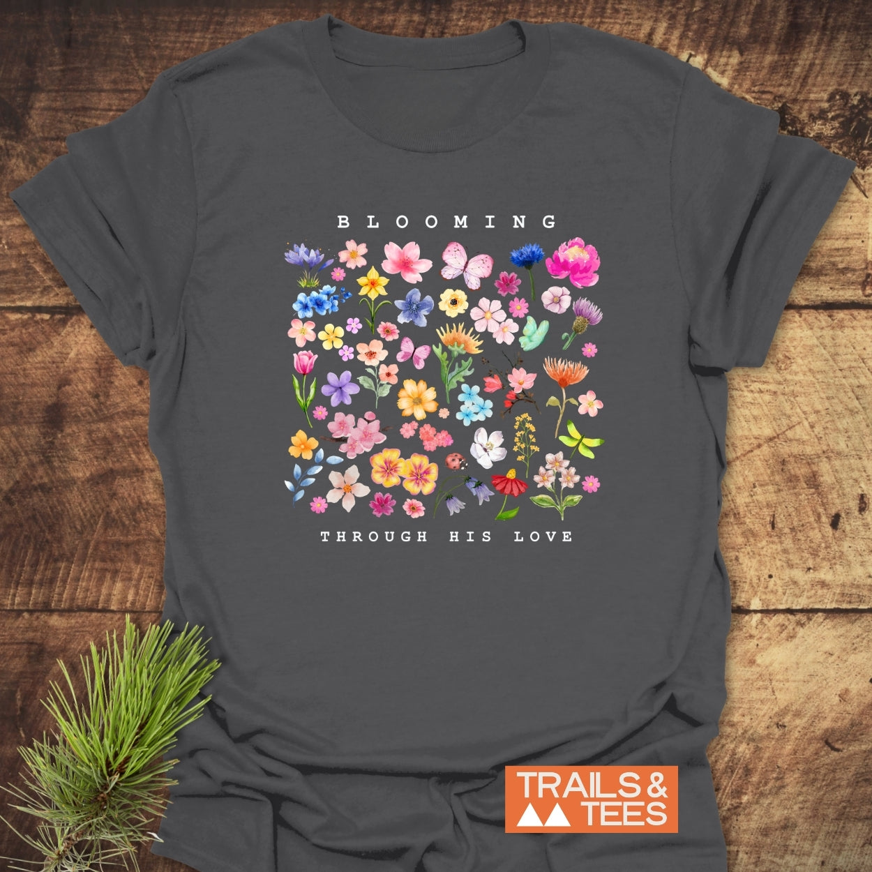 Blooming Through His Love T-Shirt featuring a colorful floral and butterfly graphic with uplifting text, laid on a wooden backdrop. Crafted from soft cotton, ideal for outdoor adventures. Logo: Trails & Tees.
