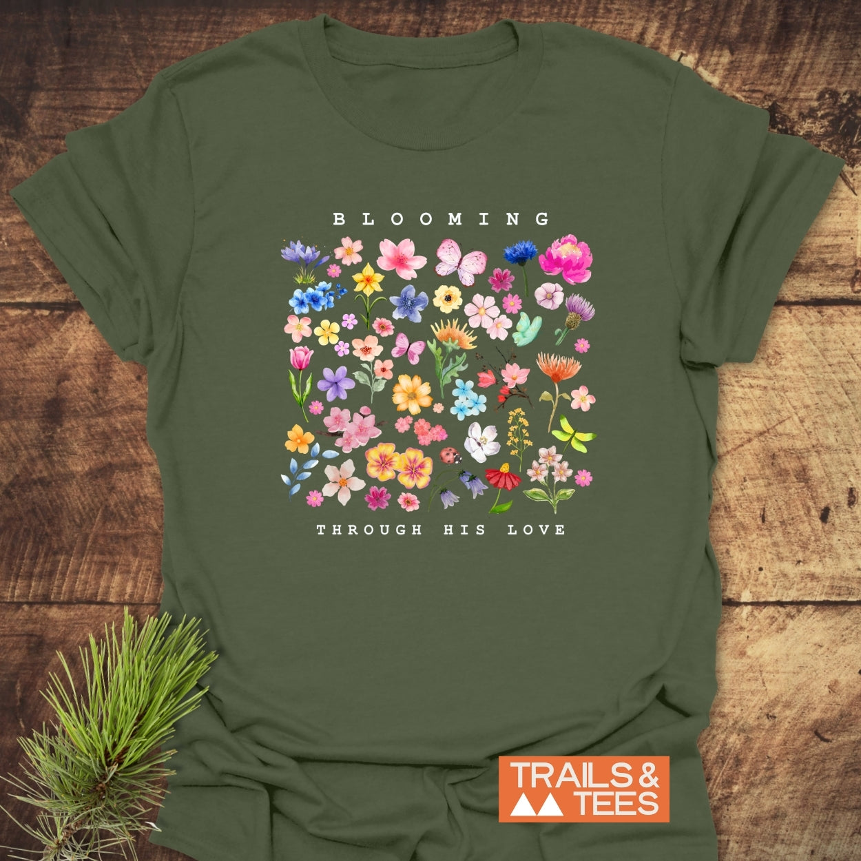 Blooming Through His Love T-Shirt flat-lay featuring watercolor flowers, butterflies, and ladybug graphic. Includes TRAILS & TEES logo badge, highlighting its comforting, durable design ideal for outdoor enthusiasts.