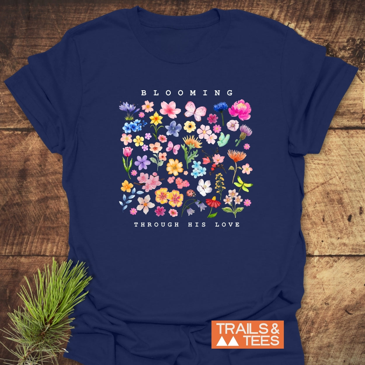Blooming Through His Love T-Shirt featuring pastel watercolor flowers in a grid with “BLOOMING” and “THROUGH HIS LOVE” text. Includes a Trails & Tees logo, laid on wood with pine greenery.