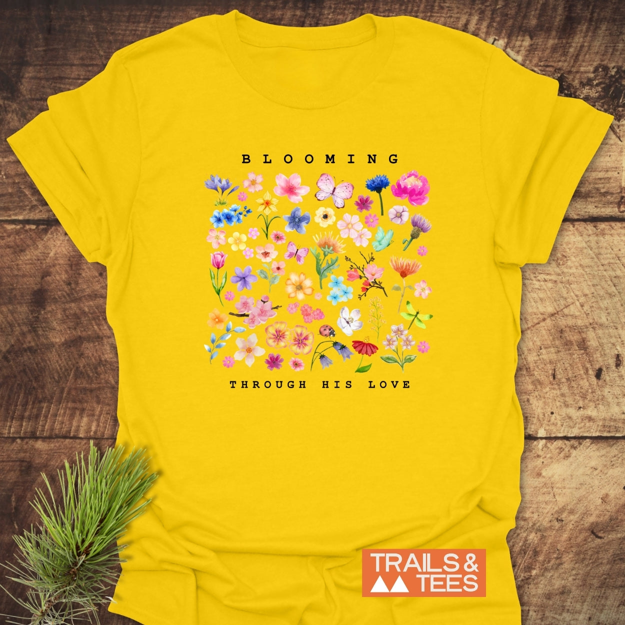 Blooming Through His Love T-Shirt featuring watercolor flowers and a butterfly, with “BLOOMING” above and “THROUGH HIS LOVE” below. Made from soft ring-spun cotton with a “TRAILS & TEES” logo.