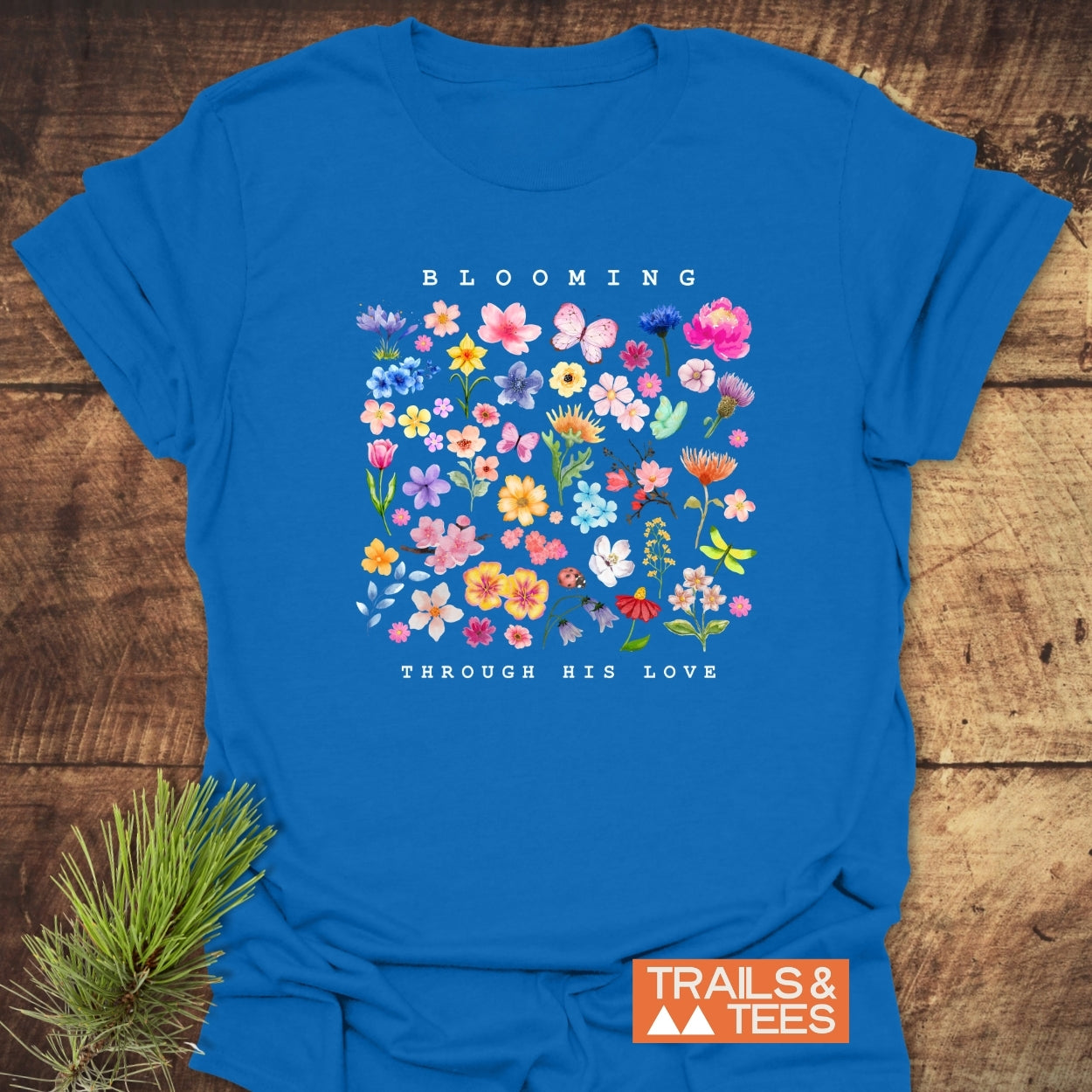 Blooming Through His Love T-Shirt featuring vibrant botanical illustrations and text, laid on a wooden surface with a pine sprig, showcasing Trails & Tees logo for an adventurous, nature-inspired look.