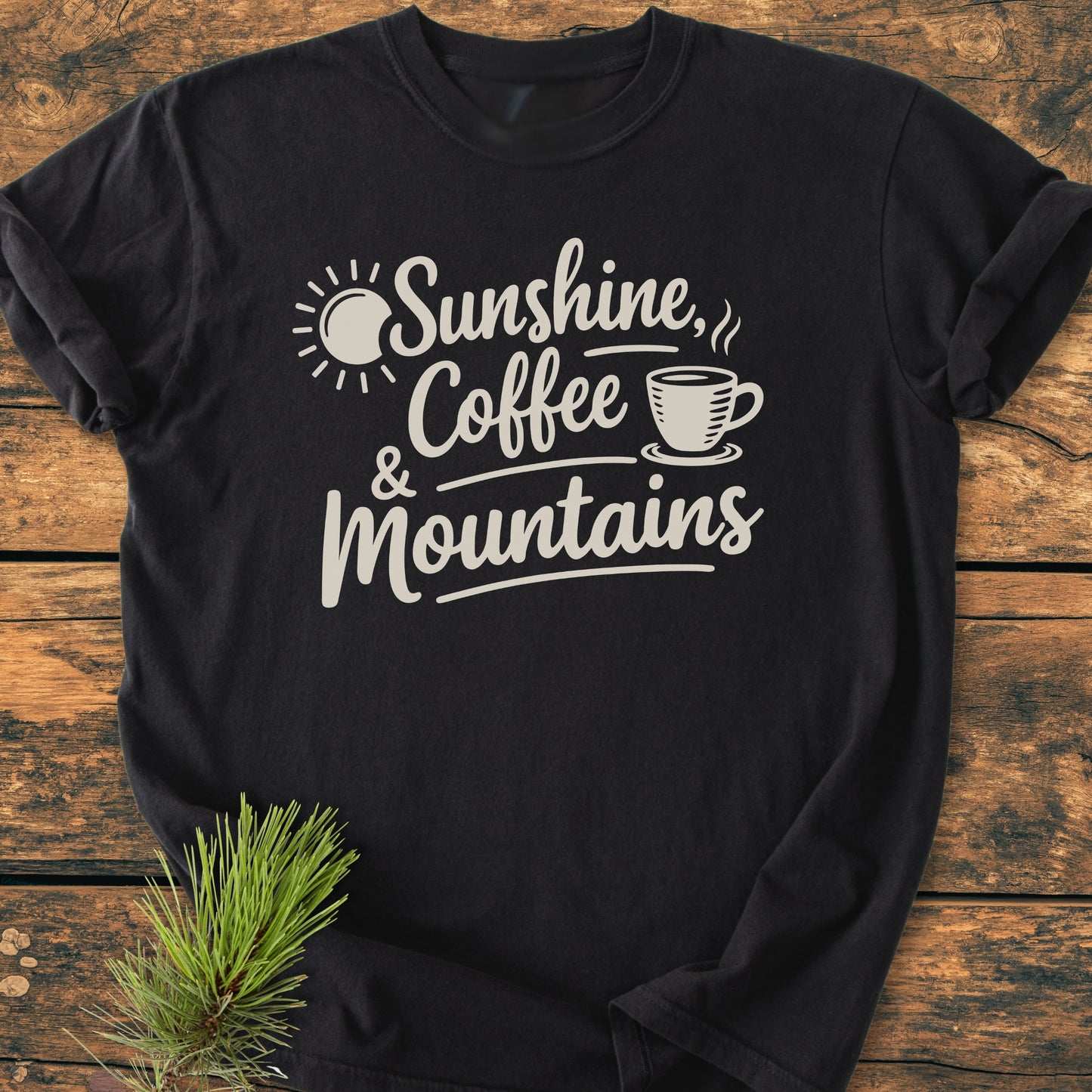 Sunshine, Coffee, Mountains - Vintage Heavyweight Tee features light-cream script on a black T-shirt, laid on rustic wood with a pine sprig, showcasing sun and coffee cup illustrations.