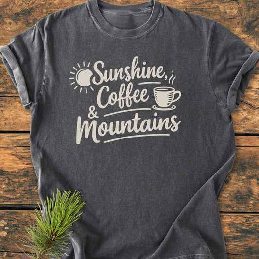 Sunshine, Coffee, Mountains - Vintage Heavyweight Tee featuring rolled sleeves, hand-lettered white graphic, and pine sprig accent, highlighting its relaxed fit and durable construction on a wooden surface.