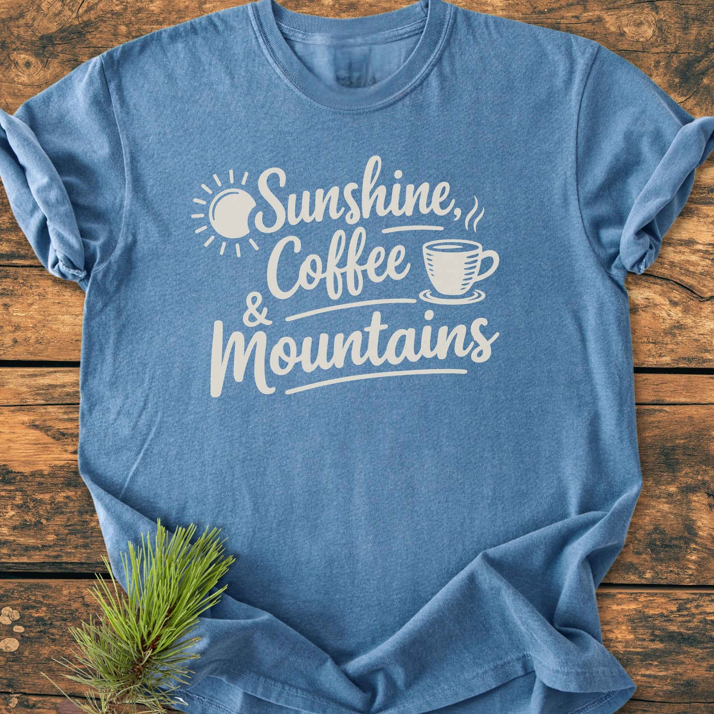 Sunshine, Coffee, Mountains - Vintage Heavyweight Tee featuring a playful script with sun and coffee icons, laid flat on wood. Includes evergreen sprig, highlighting its outdoorsy appeal and comfortable, durable design.